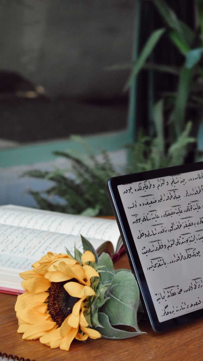 A tablet displaying Arabic text beside an open book and sunflower indoors - still life.